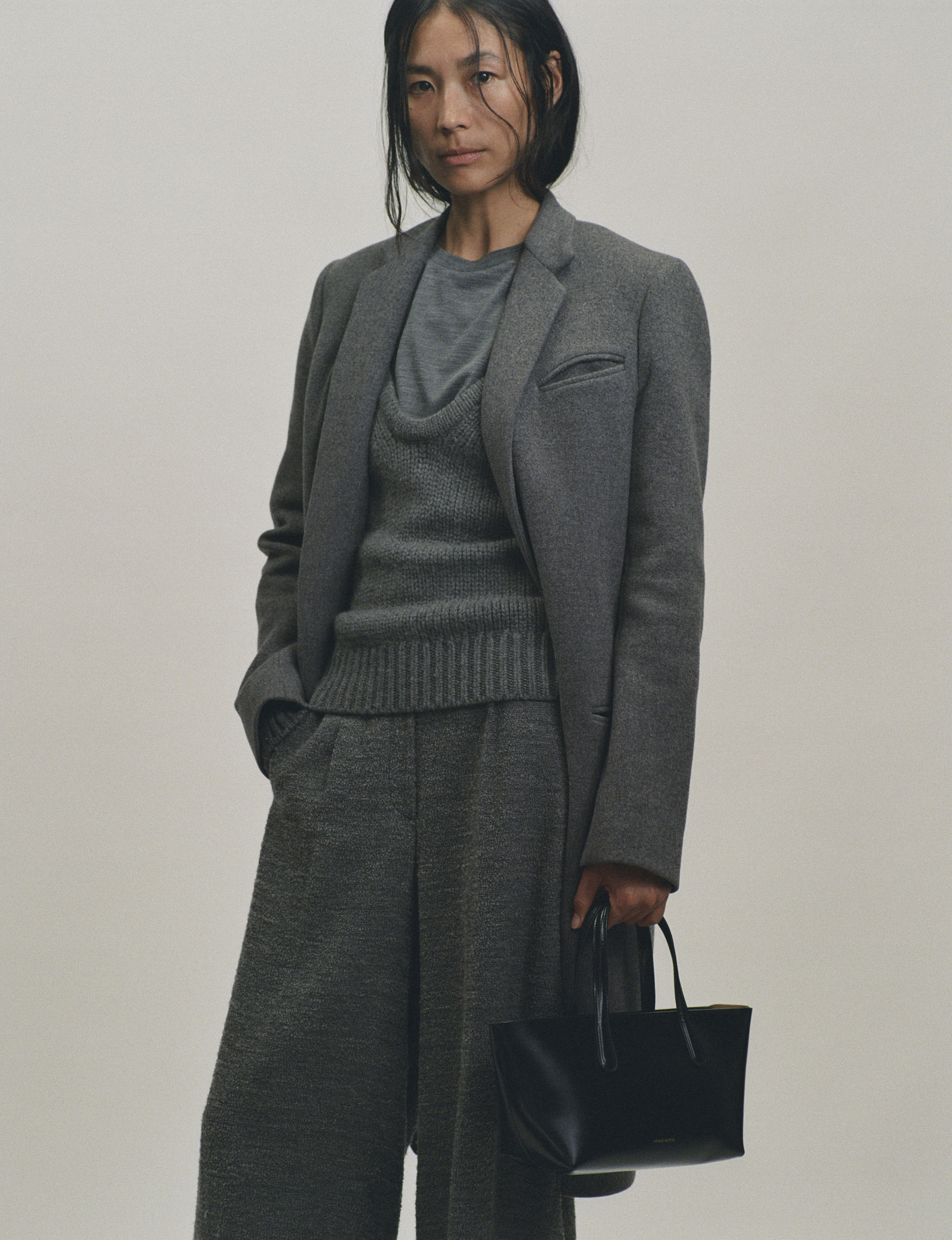 Woman in grey coat and trousers holding a black handbag against a neutral background