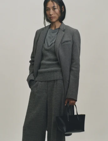 Woman in grey coat and trousers holding a black handbag against a neutral background