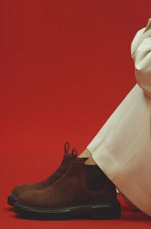 Close-up of brown suede Chelsea boots and white trousers against a vibrant red backdrop for the Mansur Gavriel fall campaign.