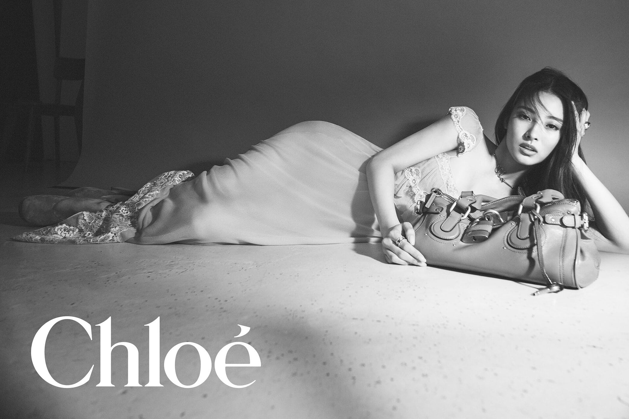 Monochrome fashion campaign image featuring a model lying beside a leather handbag with the Chloé logo visible in the foreground