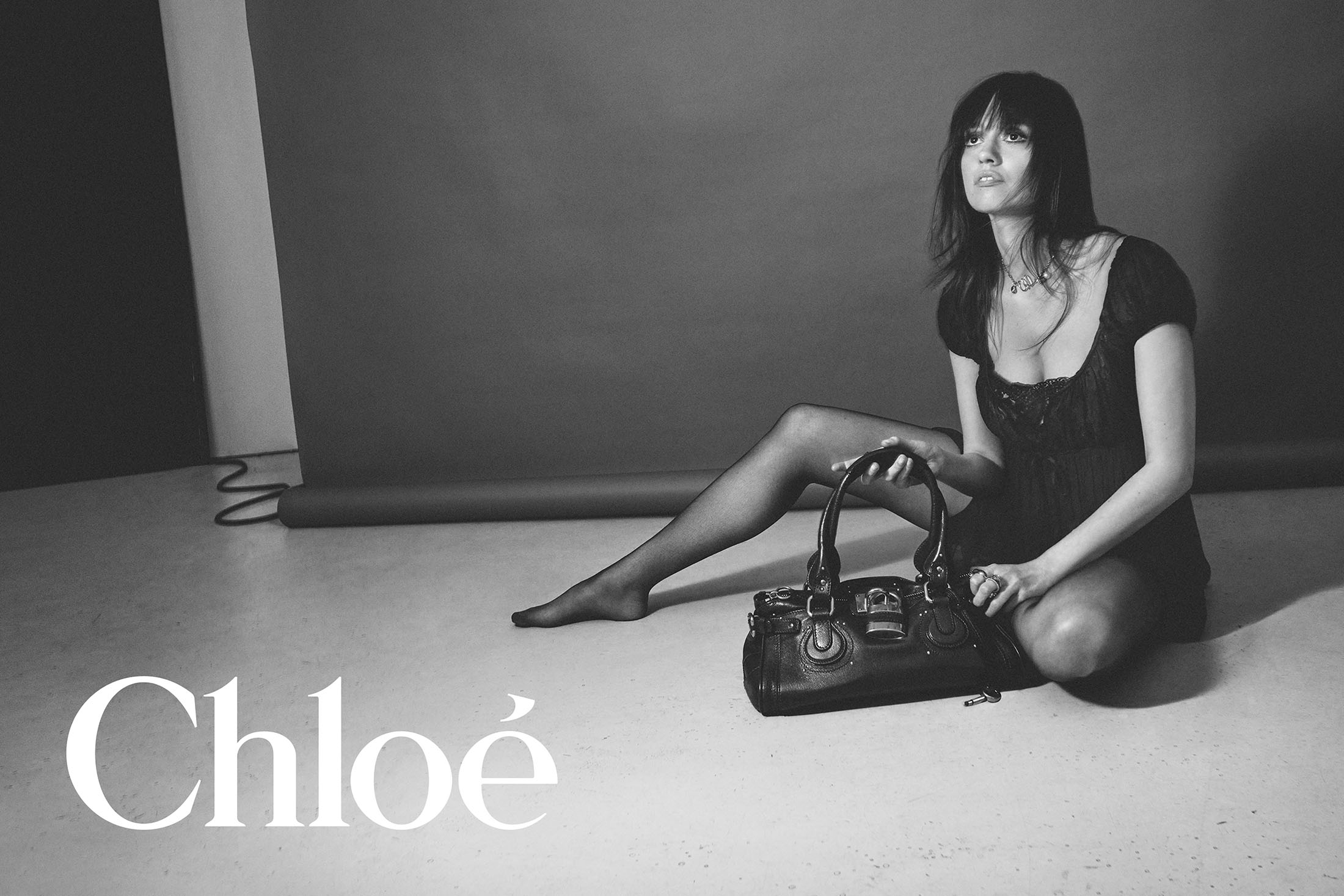 Monochrome fashion campaign image featuring a model seated on a studio floor holding a black leather handbag for Chloé.