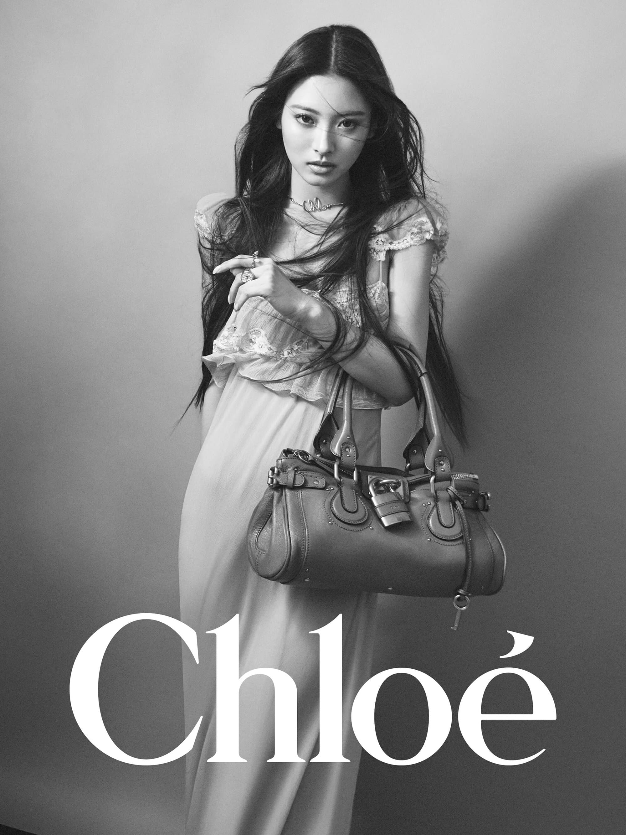 Model with long dark hair wearing a light dress and holding a leather handbag against a plain grey backdrop with Chloe text