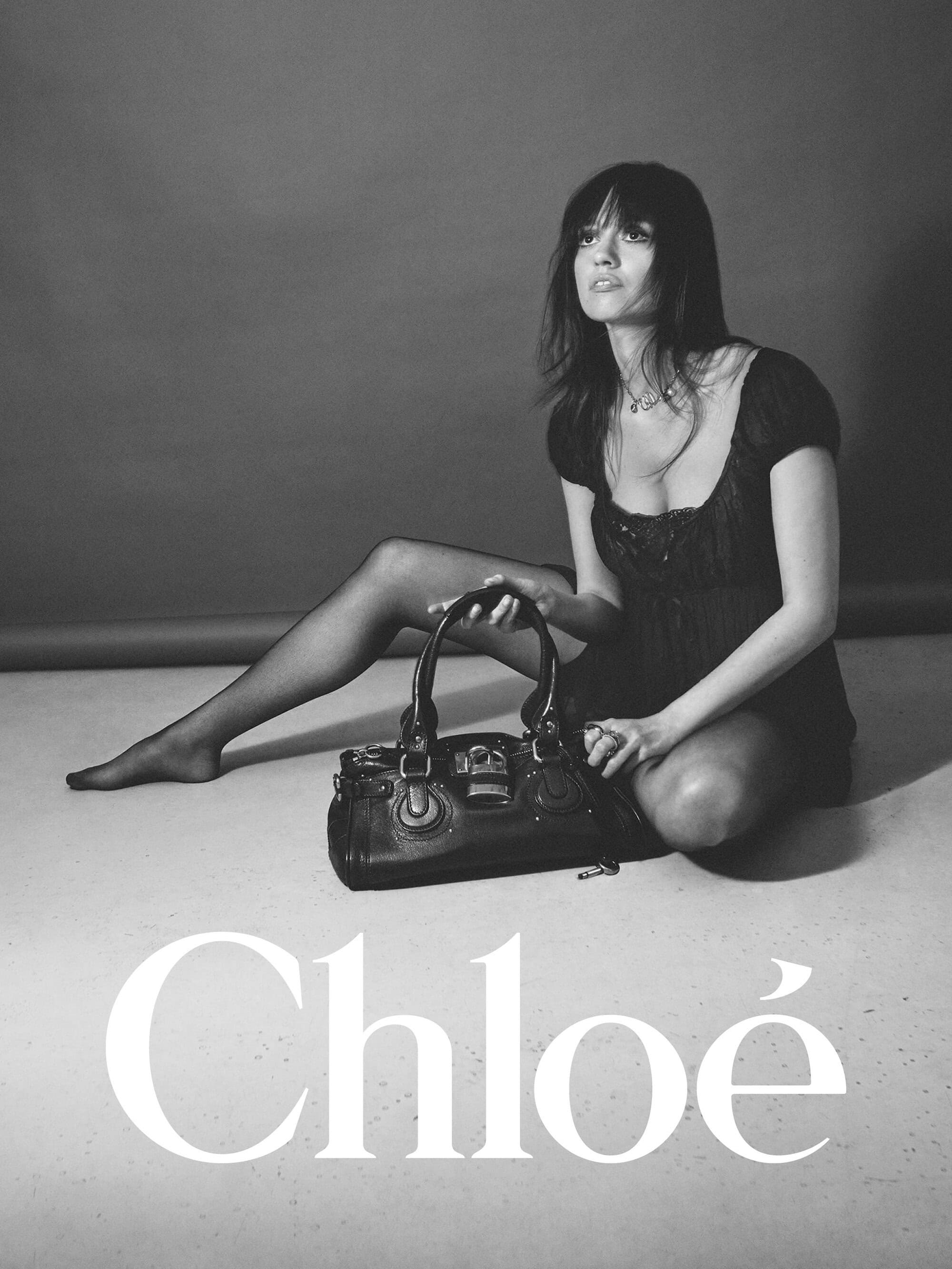 Model seated on a studio floor with a black leather handbag, wearing a dark top against a plain background