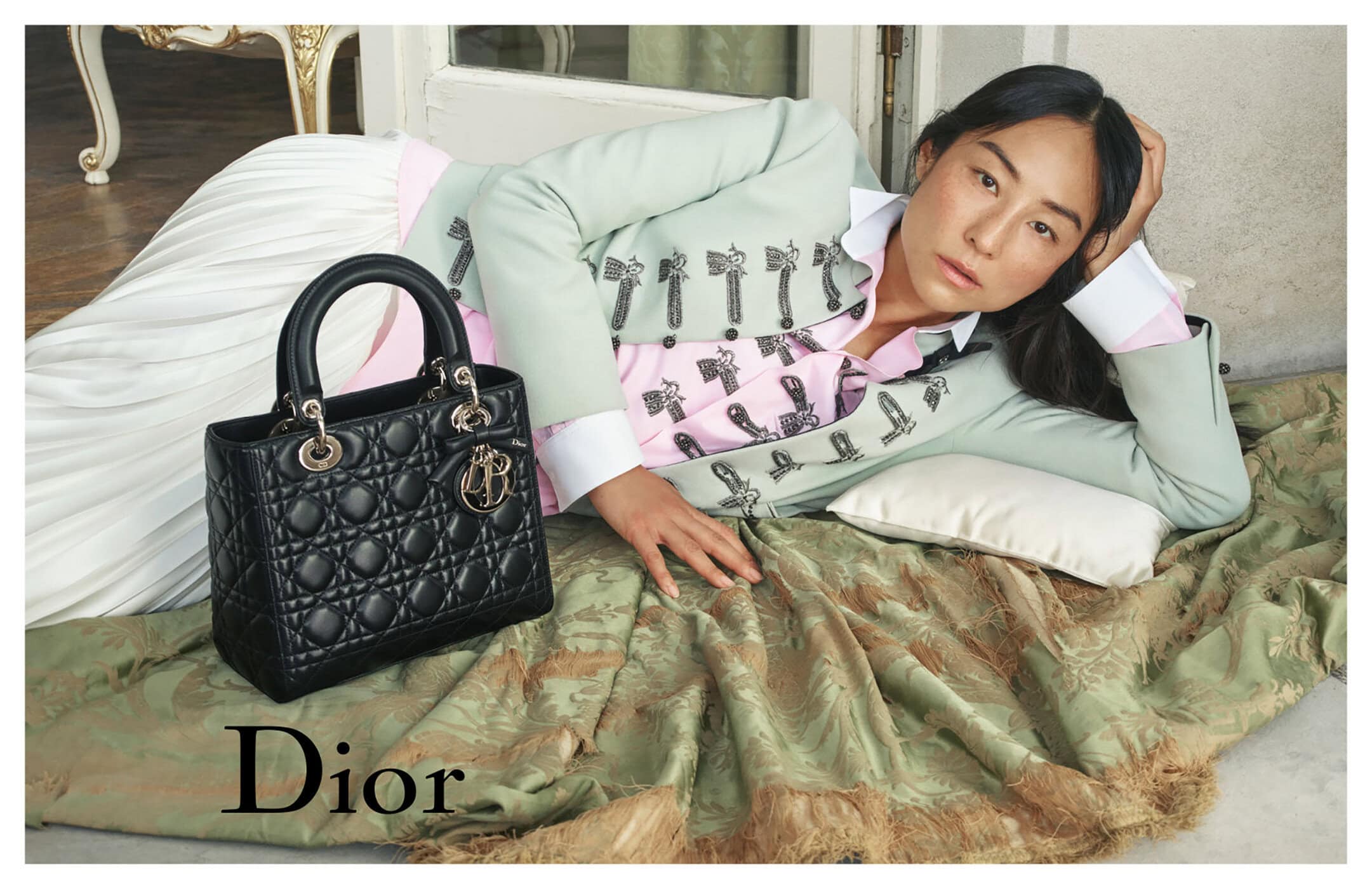 Woman in pale green jacket and white skirt reclining beside a black leather handbag for Dior ad campaign
