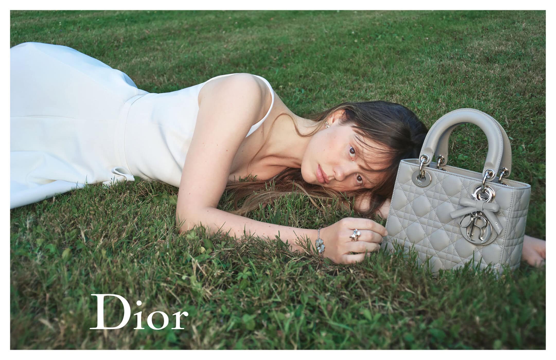 Woman in white top lying on green grass next to a grey quilted Lady Dior handbag in an outdoor ad campaign