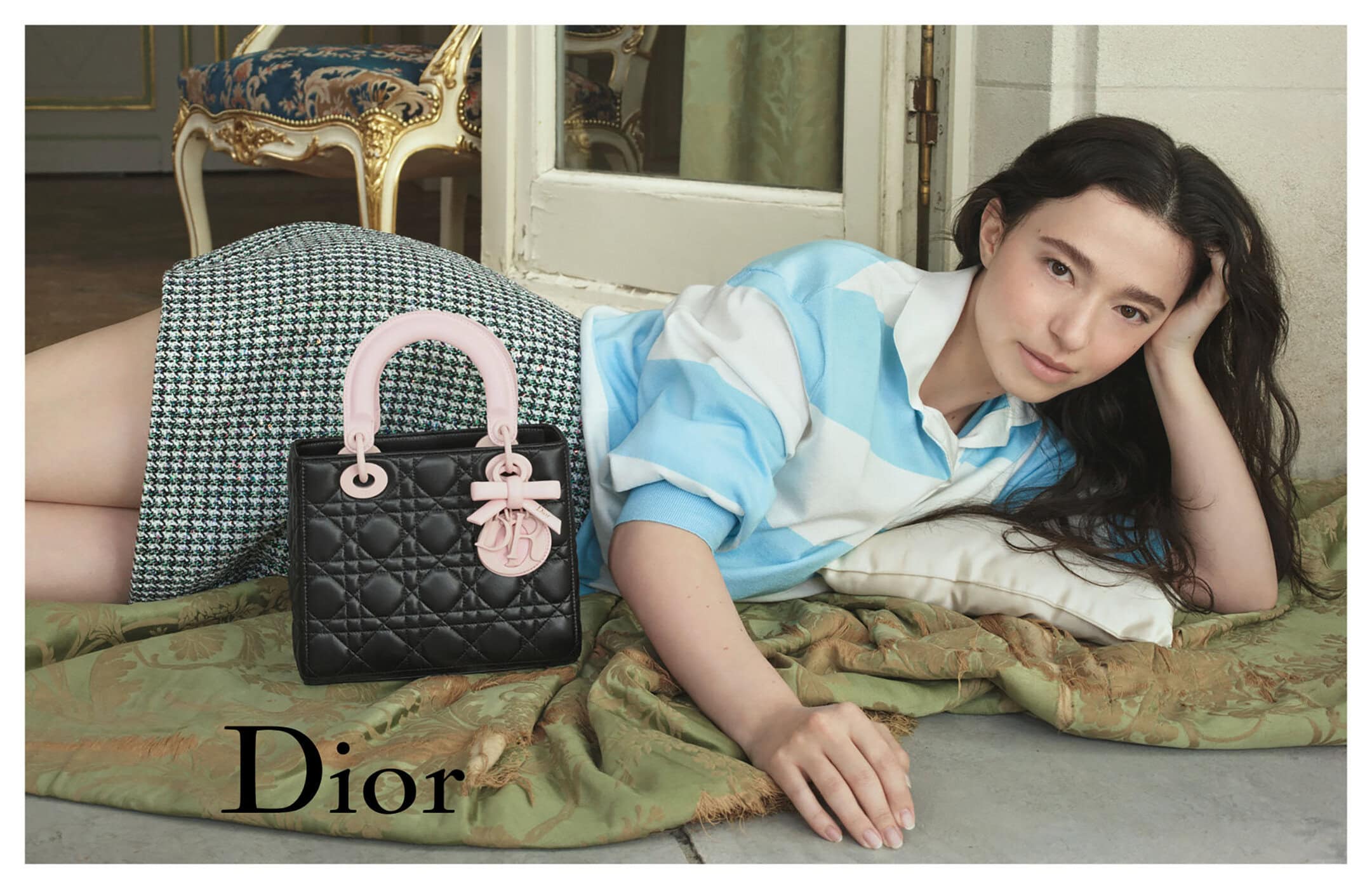 Model lying on a green patterned throw beside a black quilted leather handbag with pink handles for Dior ad campaign.