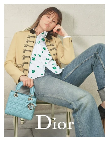 Mia Goth seated in a chair wearing a beige jacket and holding a light blue quilted handbag for Dior