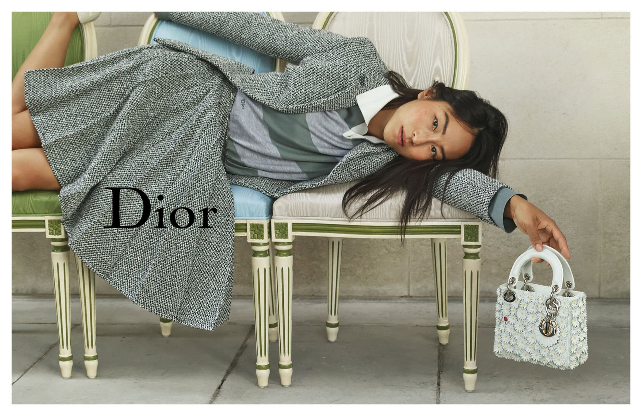 Model lying across ornate chairs wearing a grey tweed skirt suit holding a white embellished Lady Dior handbag