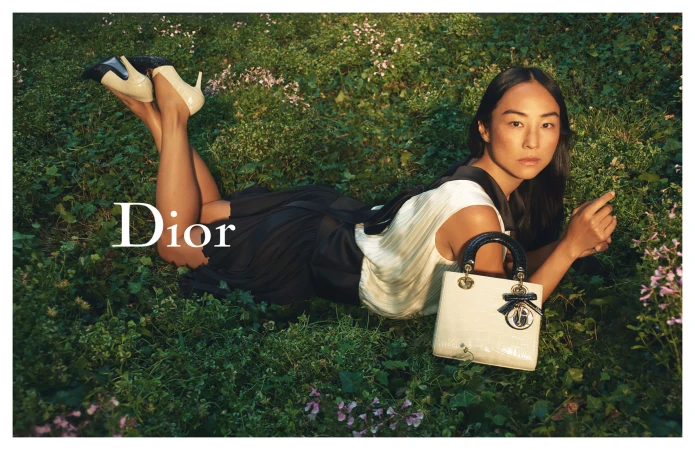 Model lying in a grassy meadow holding a white Lady Dior handbag for the 2025 ad campaign shot by David Sims