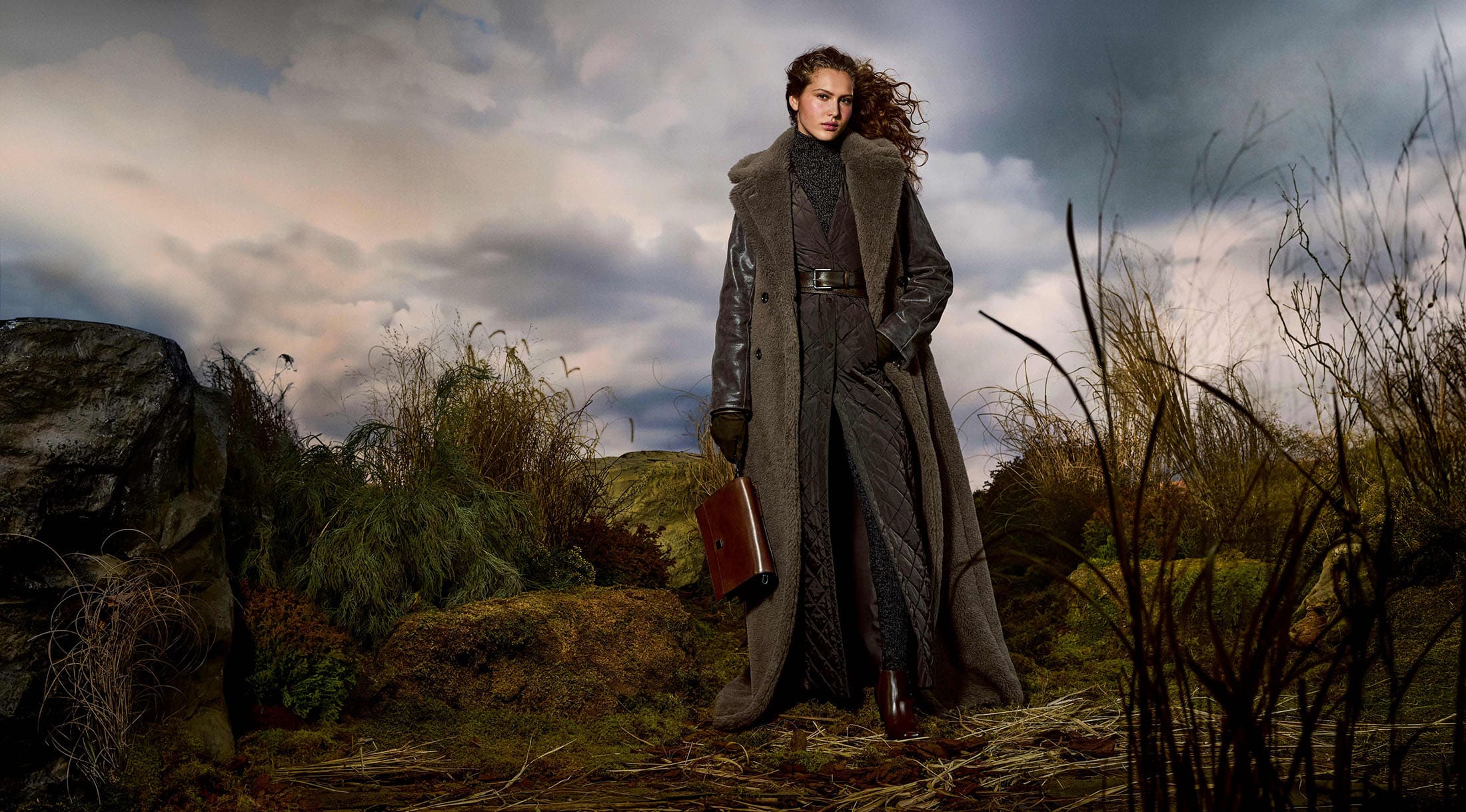 Model Angelina Kendall in a long shearling coat and holding a brown leather bag outdoors under a dramatic cloudy sky