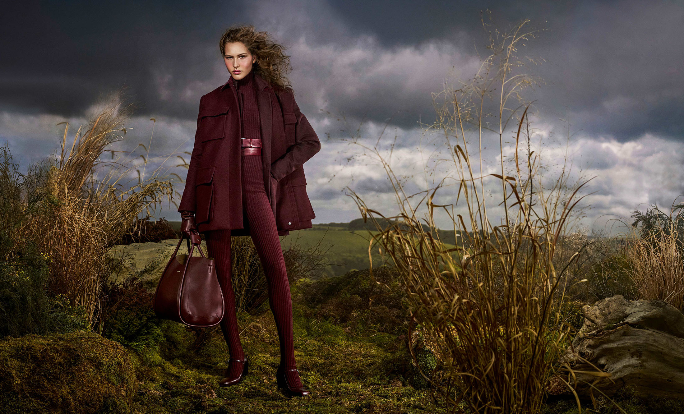 Model wearing burgundy jacket and matching knit leggings holding a leather tote bag in an overgrown landscape.