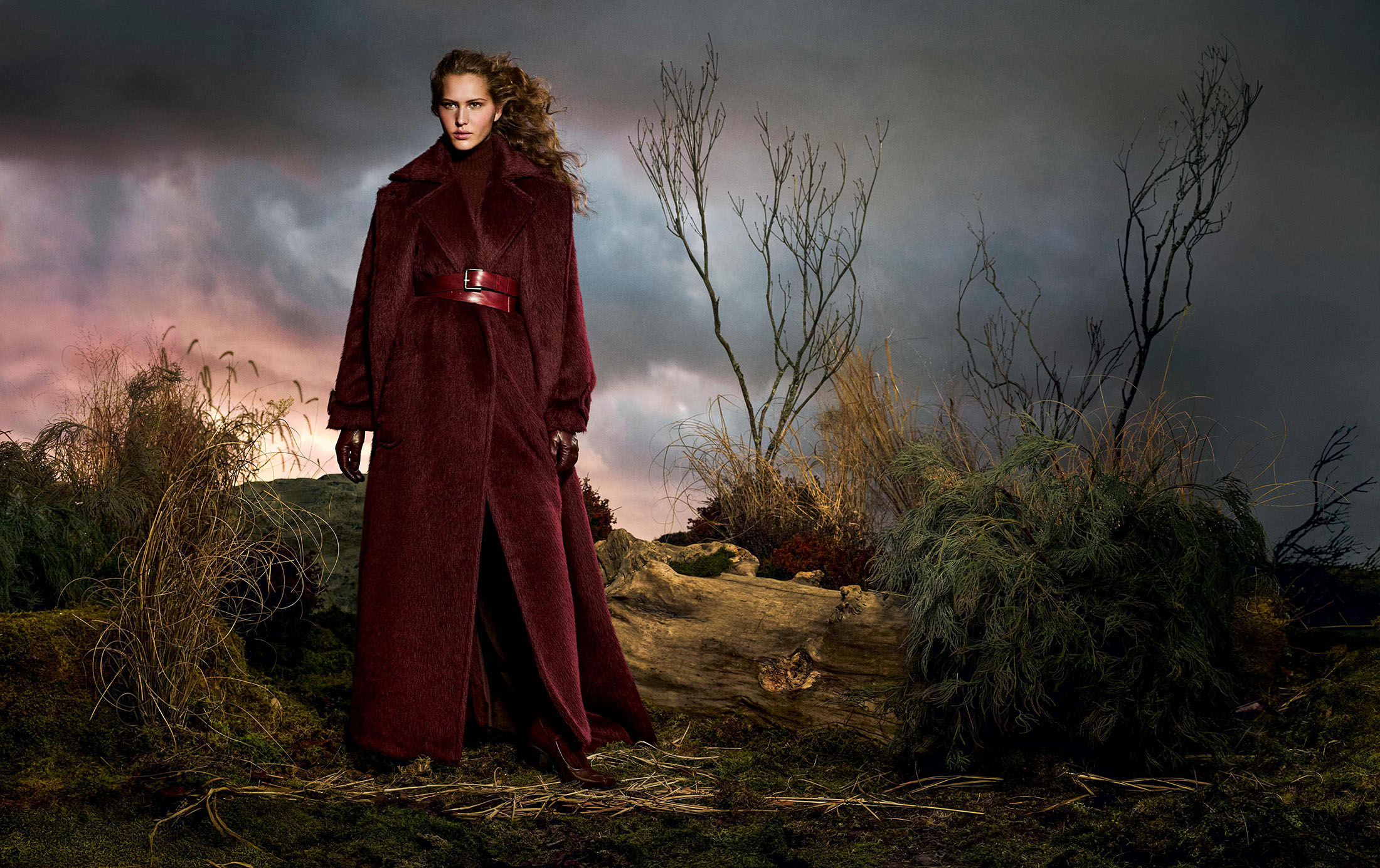 Model Angelina Kendall in a long burgundy coat standing outdoors under a dramatic stormy sky for Max Mara Fall 2025 campaign.