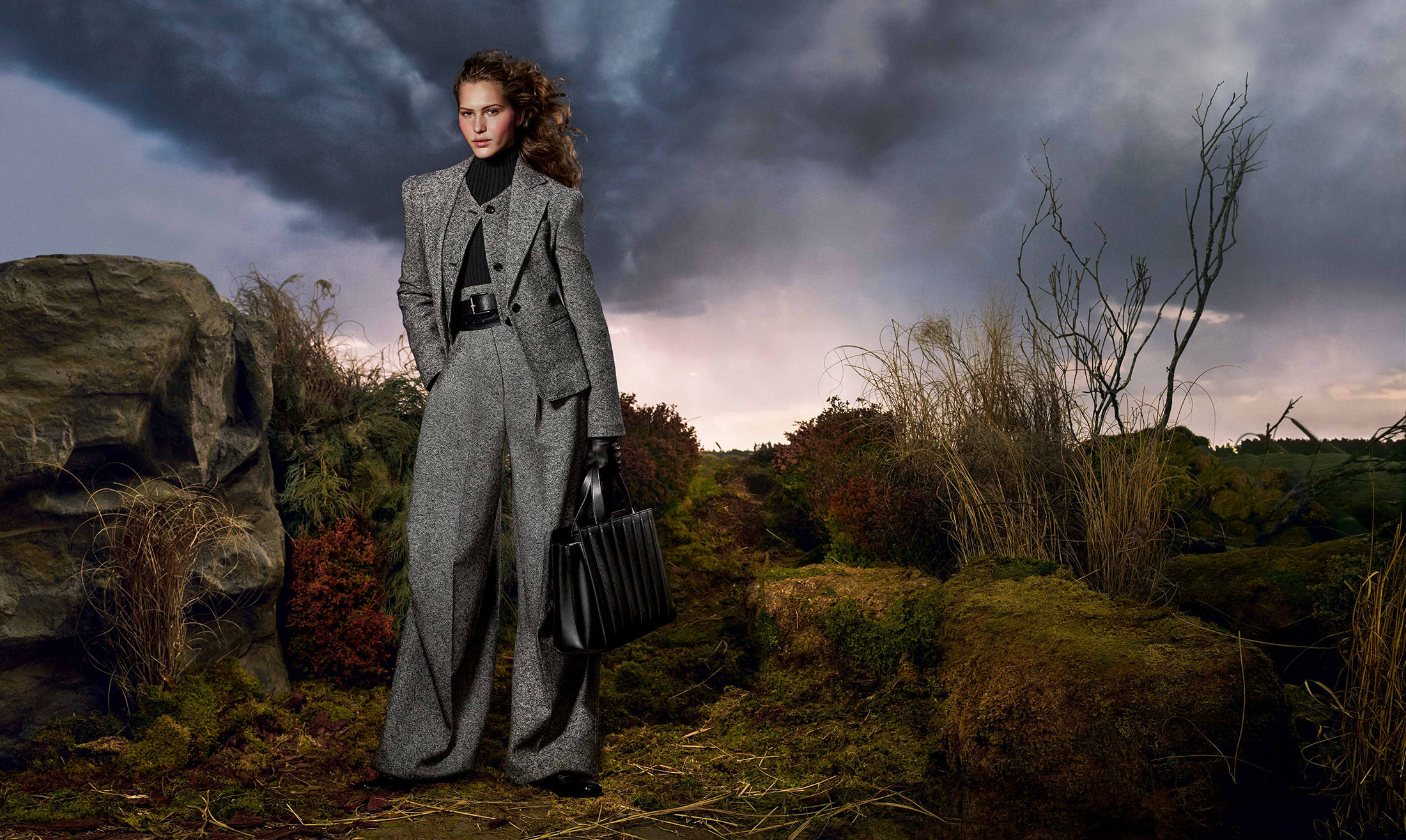 Model Angelina Kendall in a grey tweed suit holding a black leather handbag against a dramatic stormy landscape for Max Mara Fall 2025 campaign.