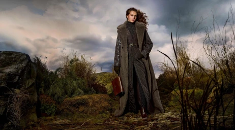 Model Angelina Kendall in a long shearling coat and holding a brown leather bag outdoors under a dramatic cloudy sky
