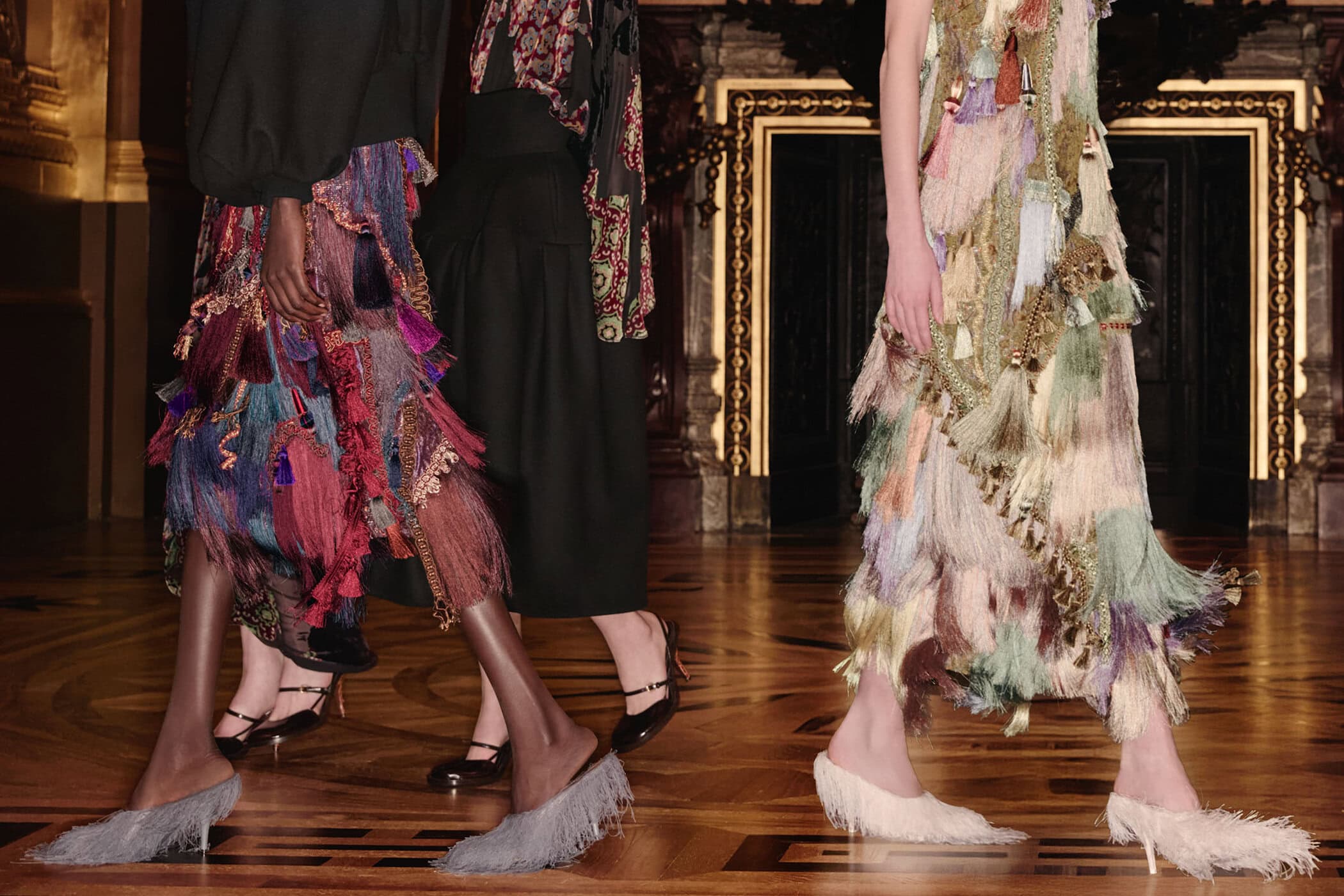 Close-up of four models in fringed skirts and ornate dresses walking on a polished wood floor inside an ornate interior.