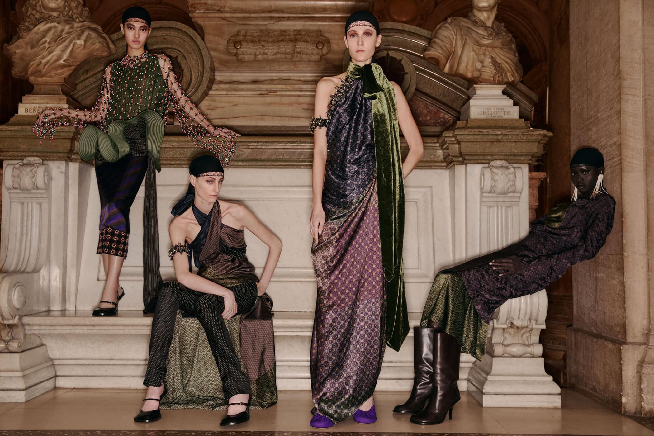 Four female models in patterned garments and head coverings posing within an interior space with classical architectural details.