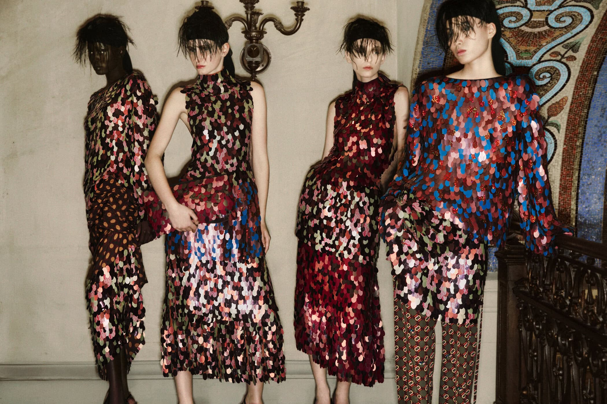 Four models in patterned sequin dresses and matching hair accessories posing indoors near an ornate wall