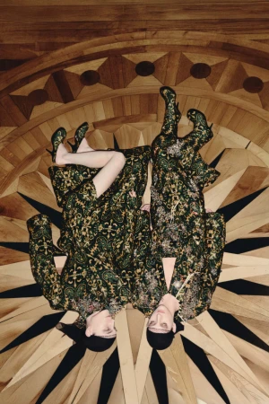 Two models wearing patterned green garments lying on an ornate geometric wooden parquet floor.