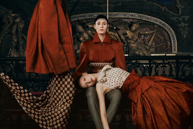 Two models in structured red attire posed within an ornate historical interior with classical sculptures.