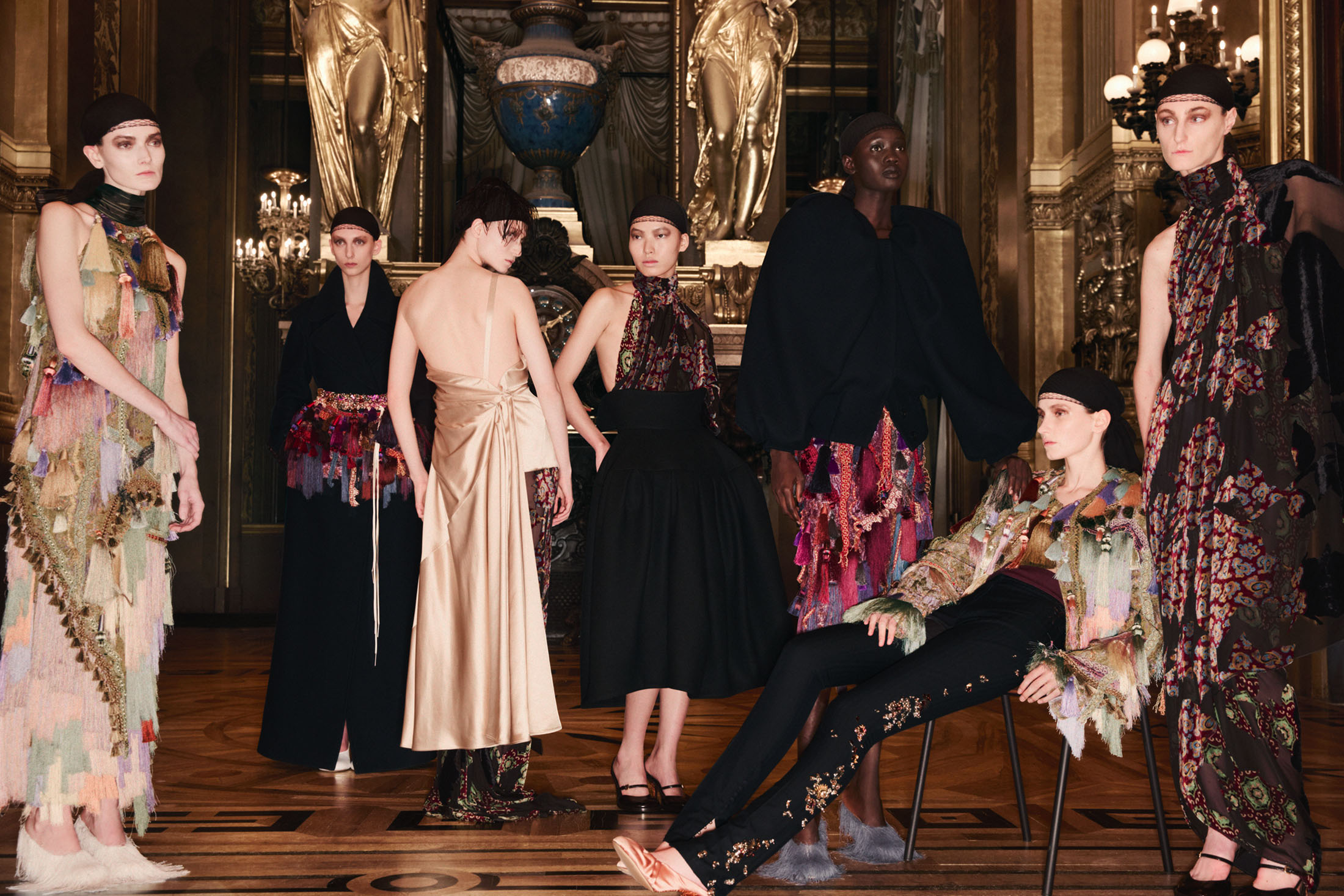 Group of models in elaborate designer fashion garments posing inside an ornate classical interior with gilded architecture.