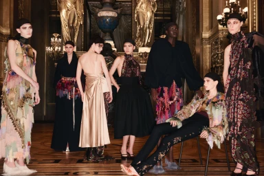Group of models in elaborate designer fashion garments posing inside an ornate classical interior with gilded architecture.