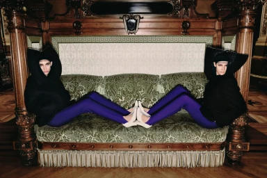 Two models wearing dark apparel and purple trousers seated facing opposite directions on an ornate green patterned sofa