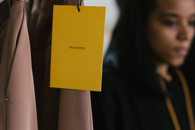Close-up of a bright yellow hanging clothing tag with black Pyer Moss text attached to brown fabric