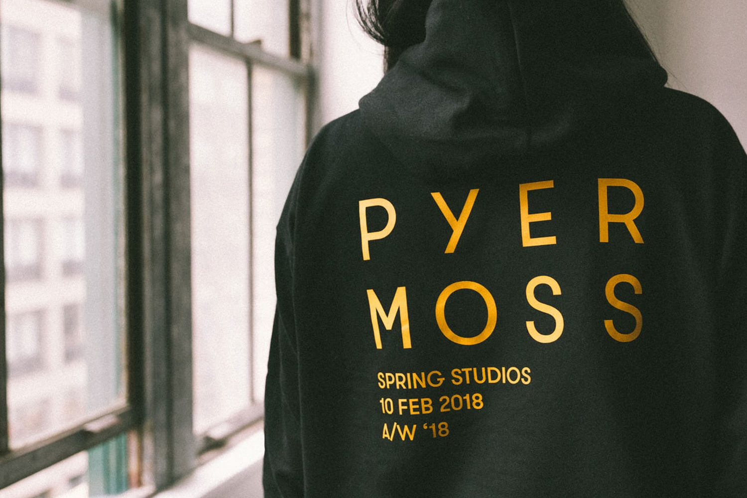 Rear view of a person wearing a black hoodie with gold lettering that reads Pyer Moss, Spring Studios, 10 Feb 2018 A/W '18.