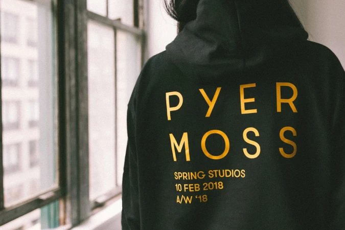 Rear view of a person wearing a black hoodie with gold lettering that reads Pyer Moss, Spring Studios, 10 Feb 2018 A/W '18.