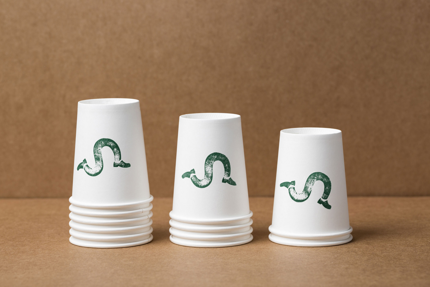 Three stacks of white paper cups with green printed worm logos arranged on a brown textured background.