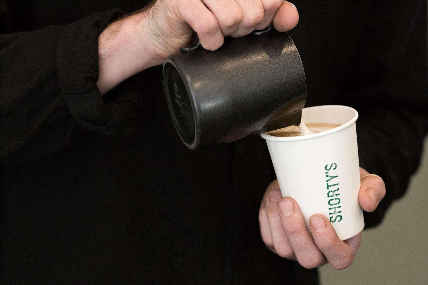 Close-up of hands pouring steamed milk from a black pitcher into a branded white paper coffee cup