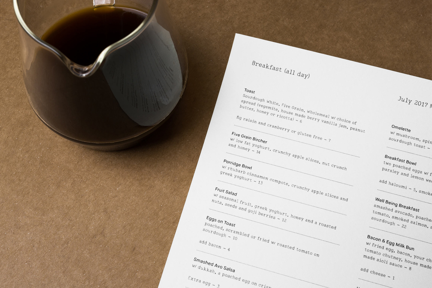Printed breakfast menu next to a glass pitcher of dark coffee on a brown textured surface