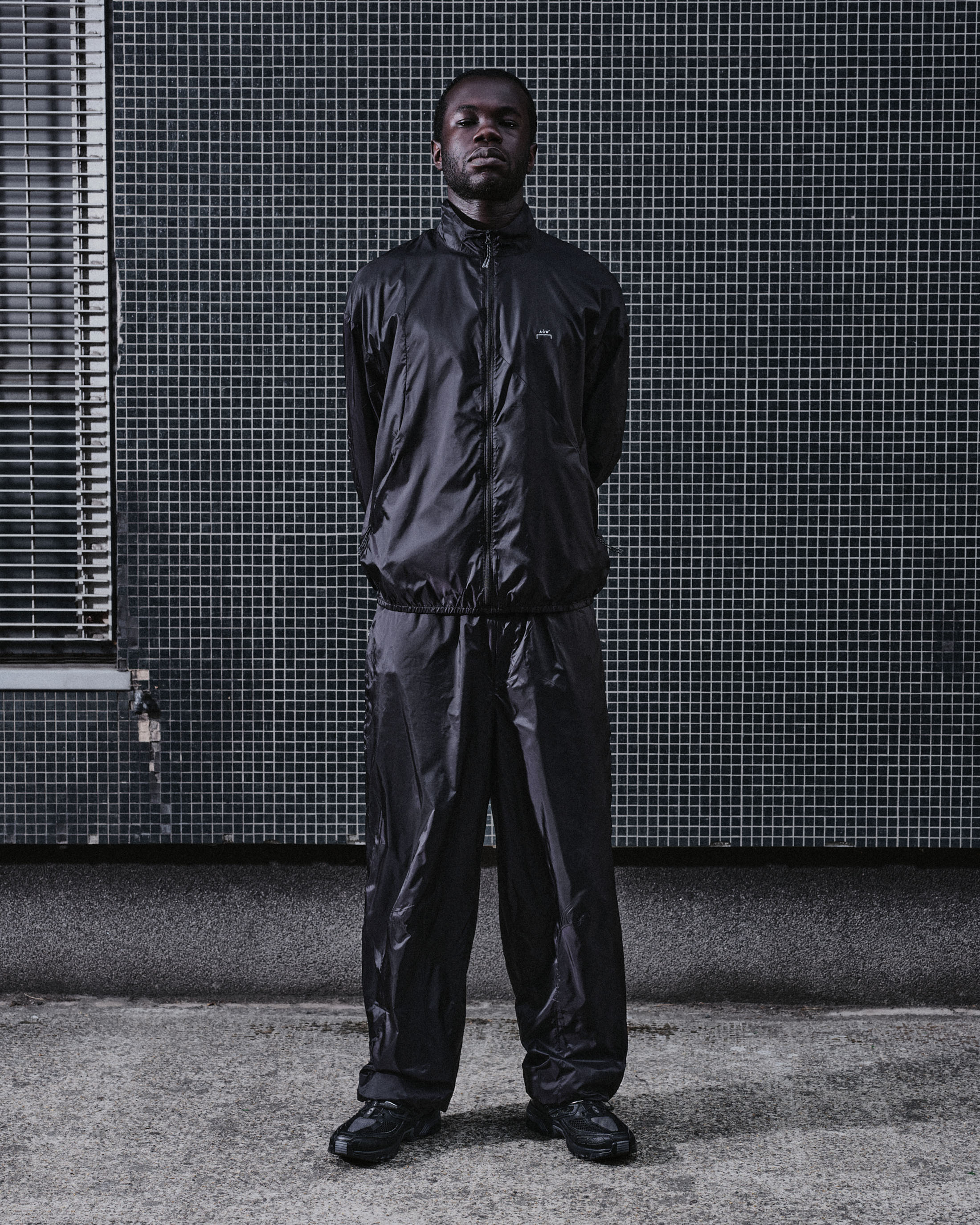 Black man standing straight against a tiled building wall wearing a dark track suit and sneakers