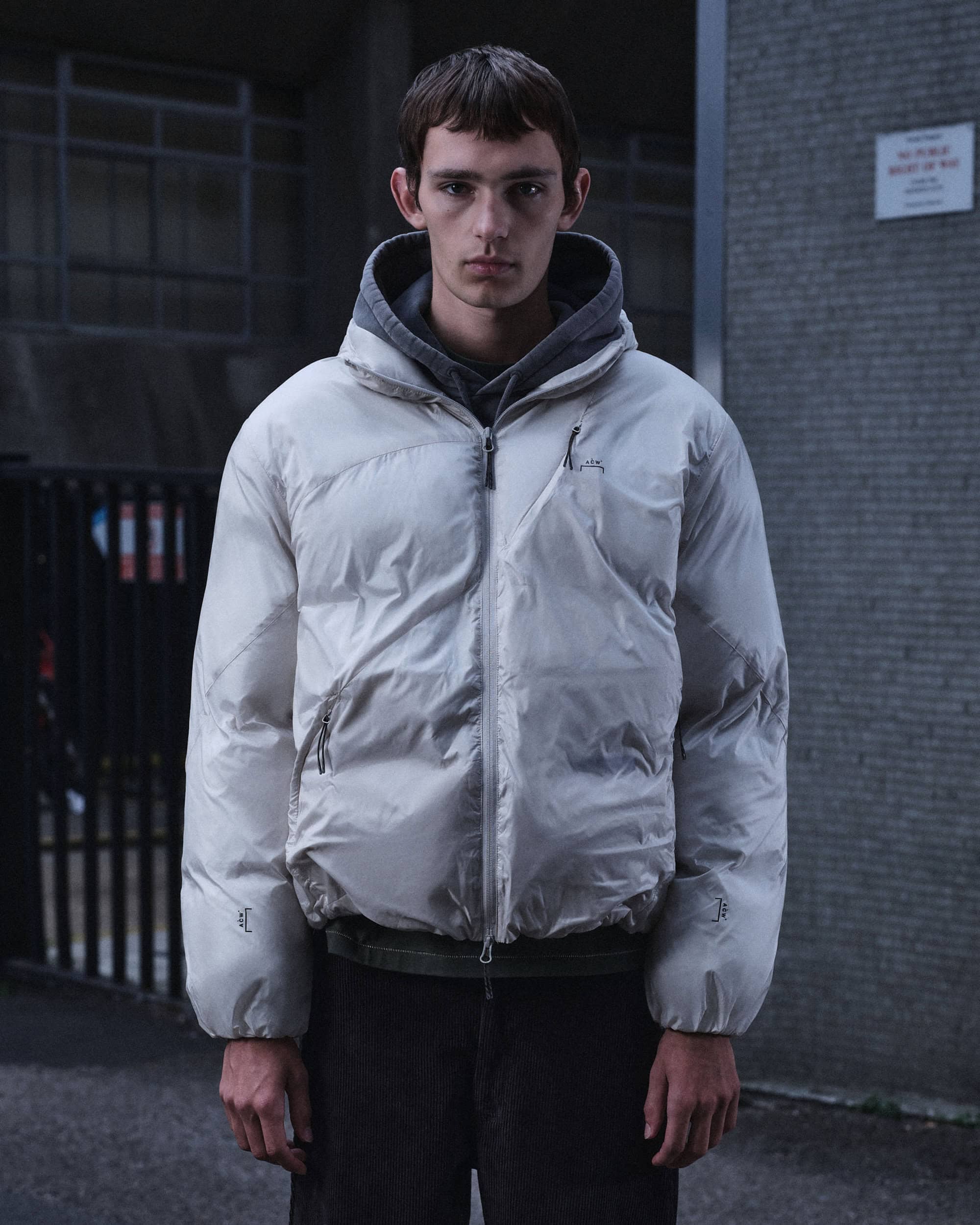 A-Cold-Wall model wearing a white puffer jacket and grey hoodie standing outdoors against a grey brick building.