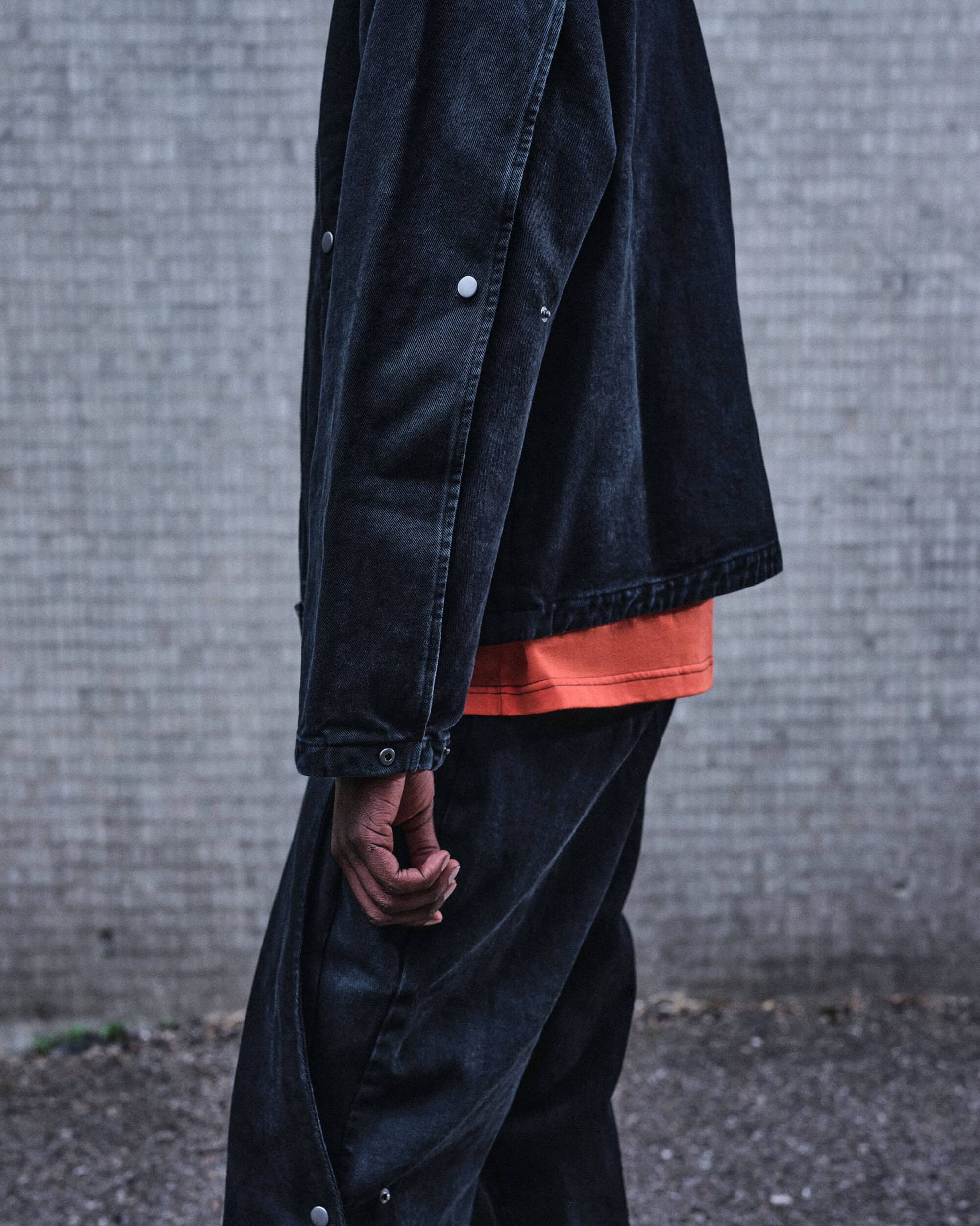 Close-up of a person wearing a dark denim jacket over an orange t-shirt and matching denim trousers against a concrete wall.