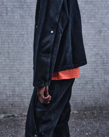 Close-up of a person wearing a dark denim jacket over an orange t-shirt and matching denim trousers against a concrete wall.