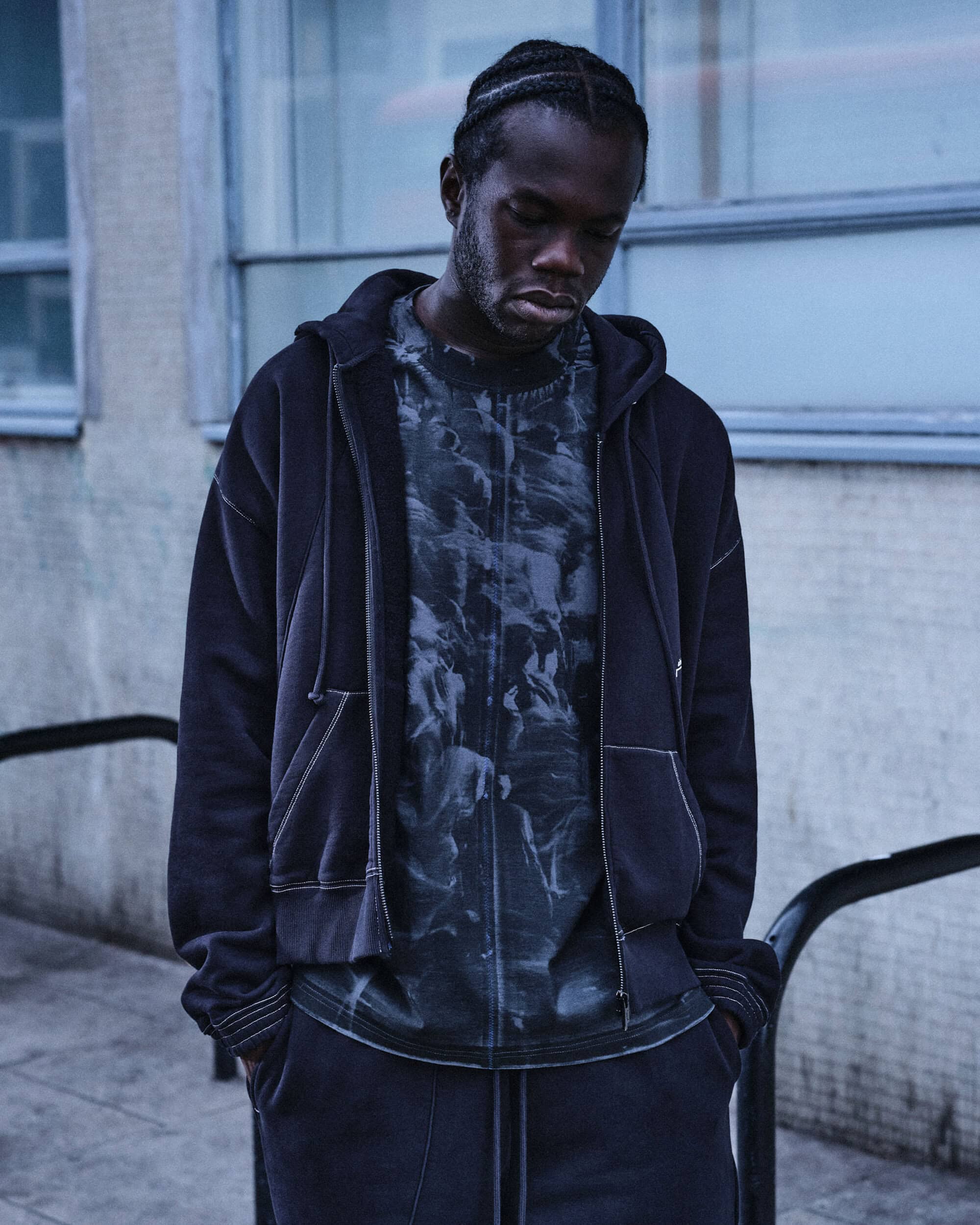Black man wearing a dark zip-up hoodie and graphic t-shirt against a concrete building exterior