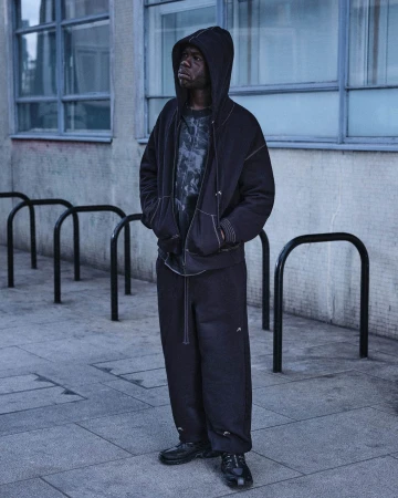 Man wearing a black hoodie and matching trousers standing outdoors against an urban architectural background.