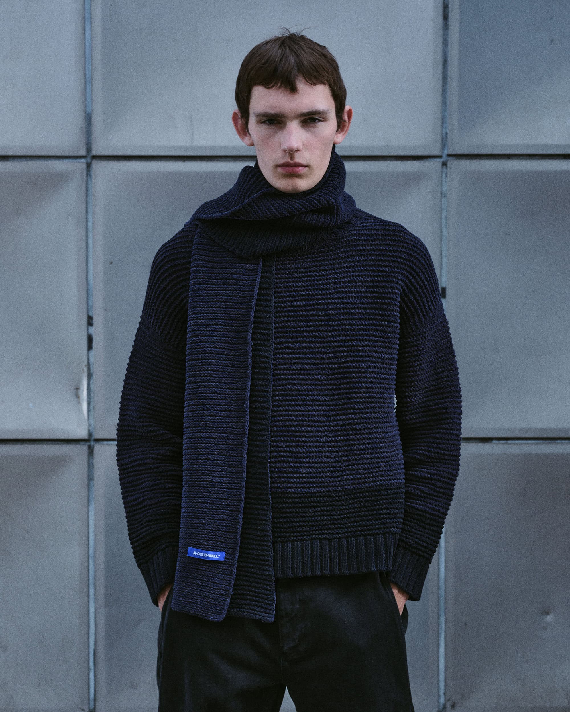 Model wearing a dark navy ribbed knit sweater and matching scarf standing before a geometric metal wall panel background.