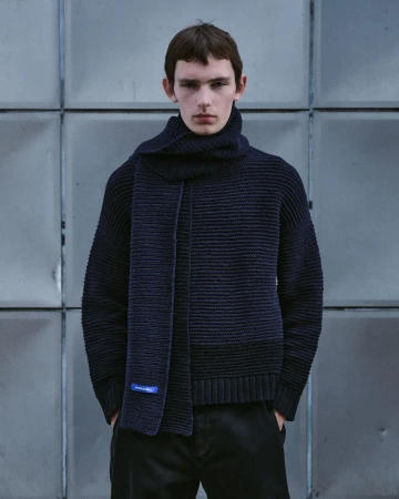 Model wearing a dark navy ribbed knit sweater and matching scarf standing before a geometric metal wall panel background.