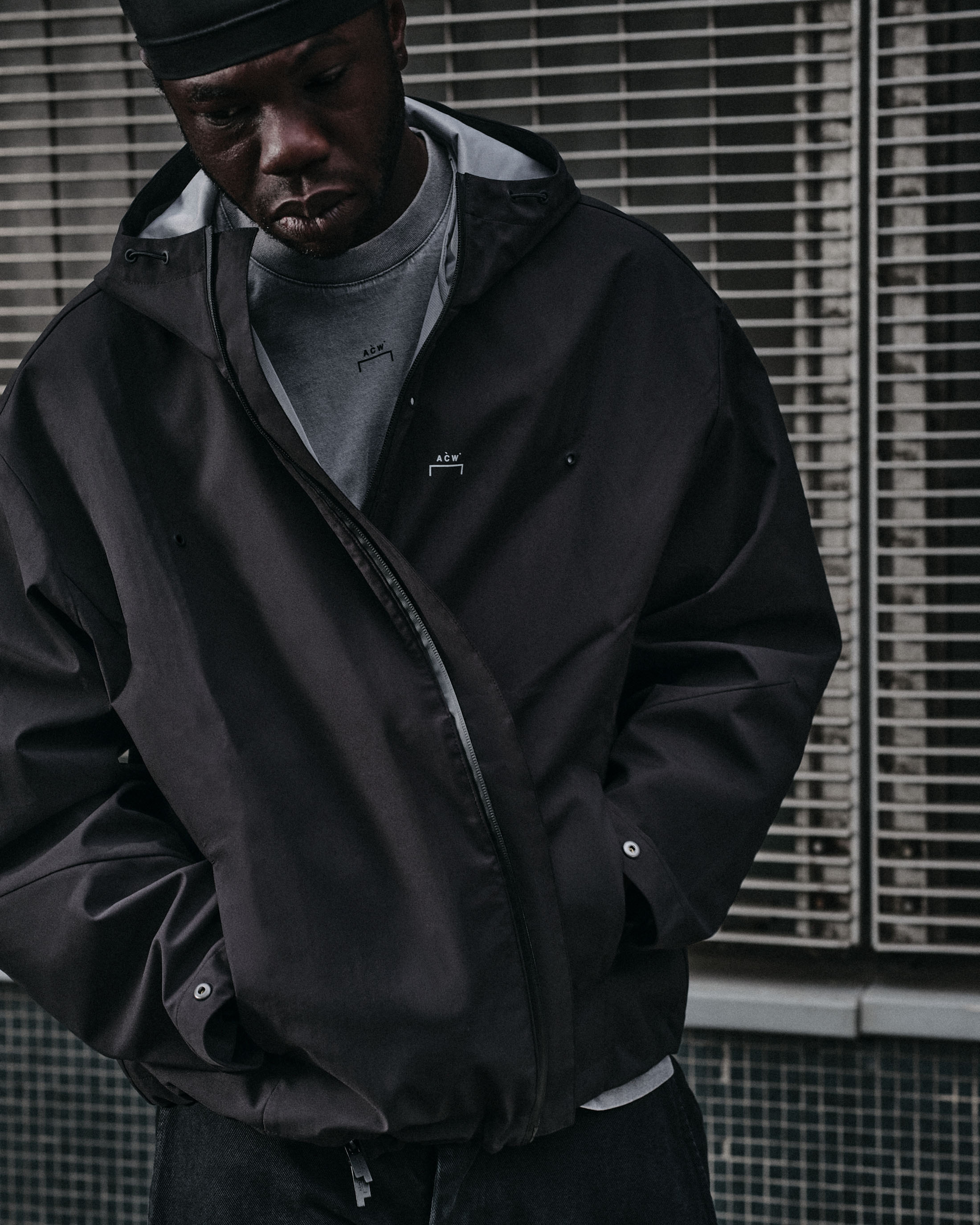 Black man wearing a black hooded jacket and grey crewneck sweater standing in front of metal grating.