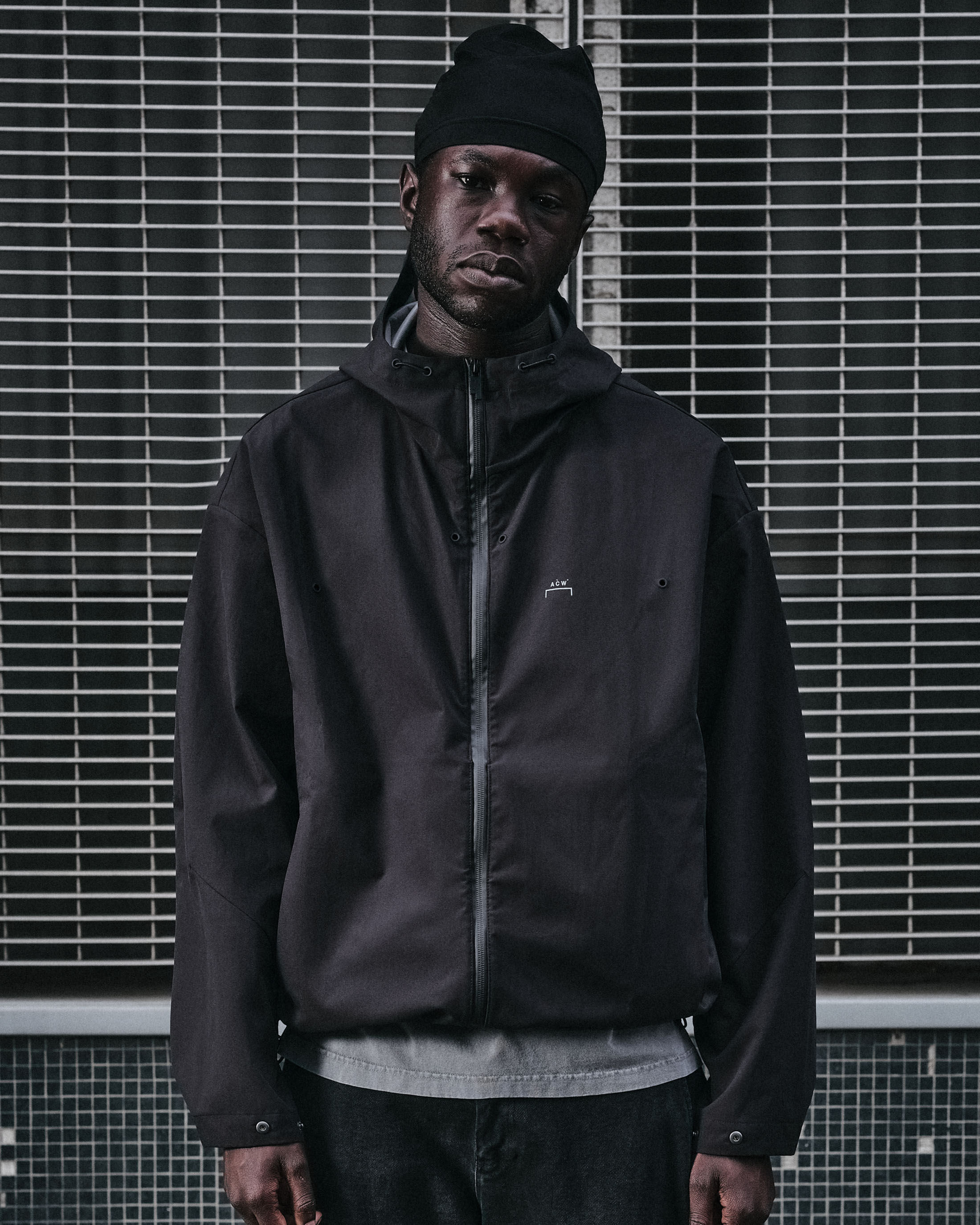 Black man wearing a dark technical jacket and beanie against a metal grille background for A-Cold-Wall