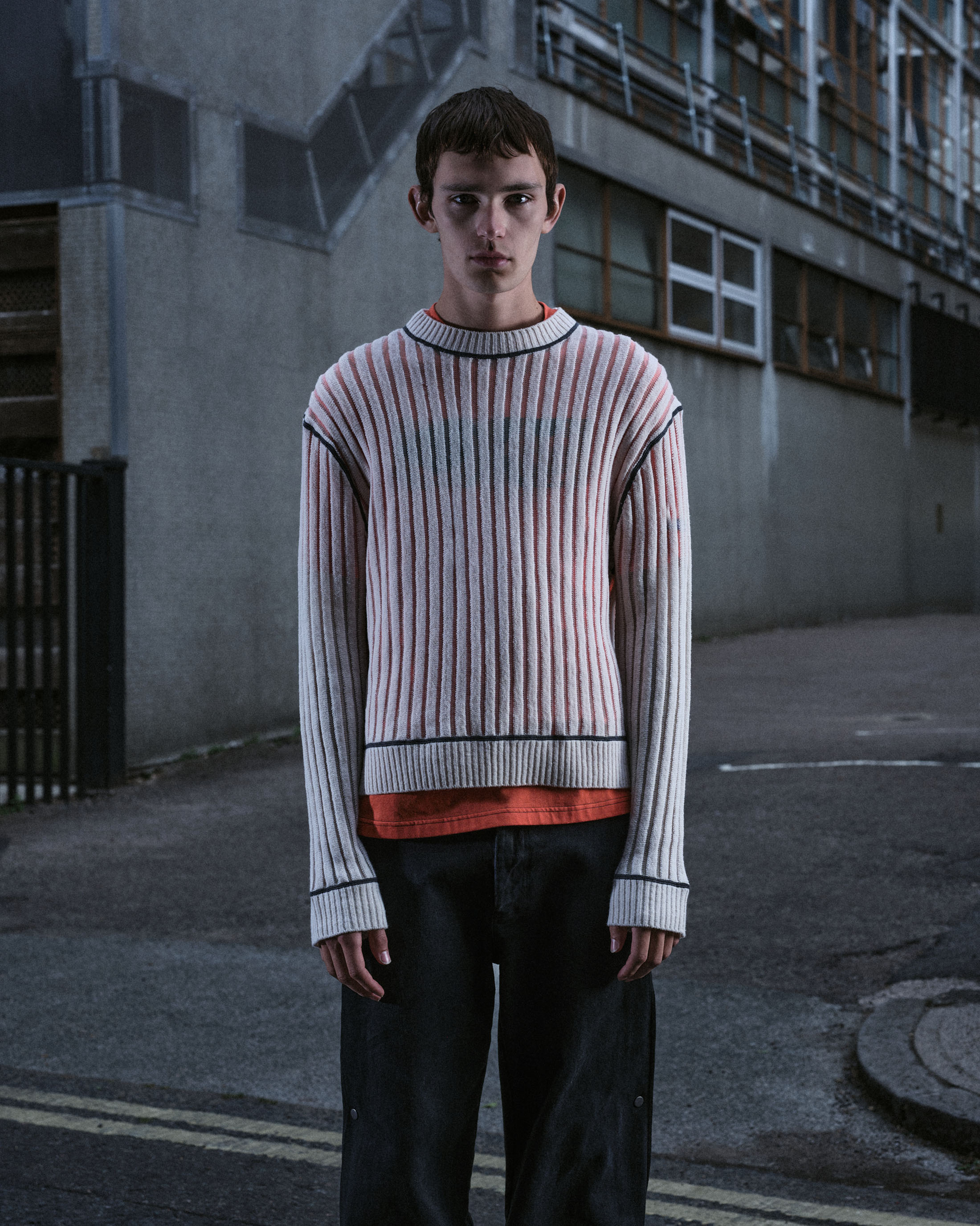 A young male model standing on an urban street wearing a ribbed white sweater over an orange t-shirt and dark trousers.