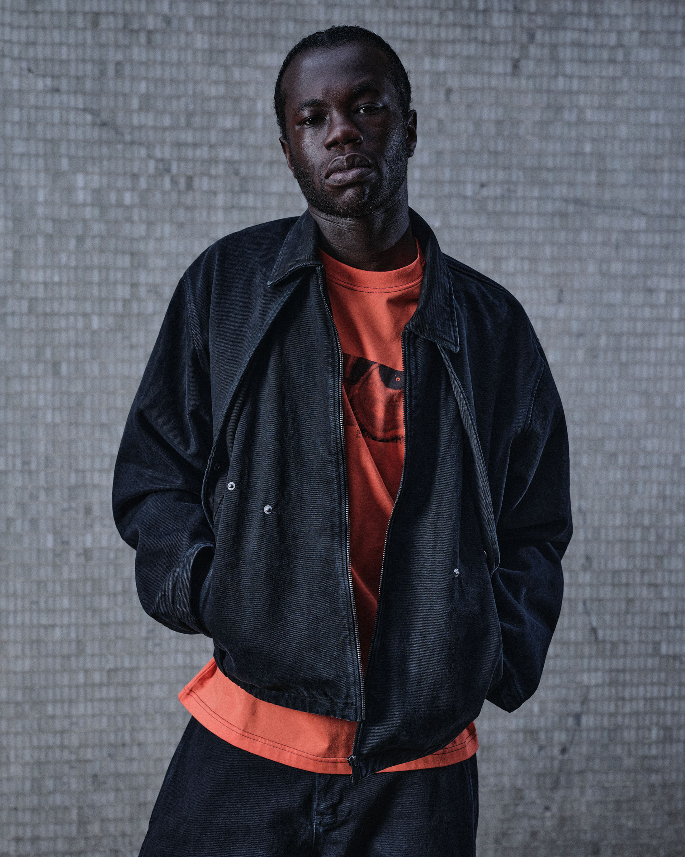 Black man wearing a dark jacket over an orange t-shirt against a textured grey wall for A-Cold-Wall