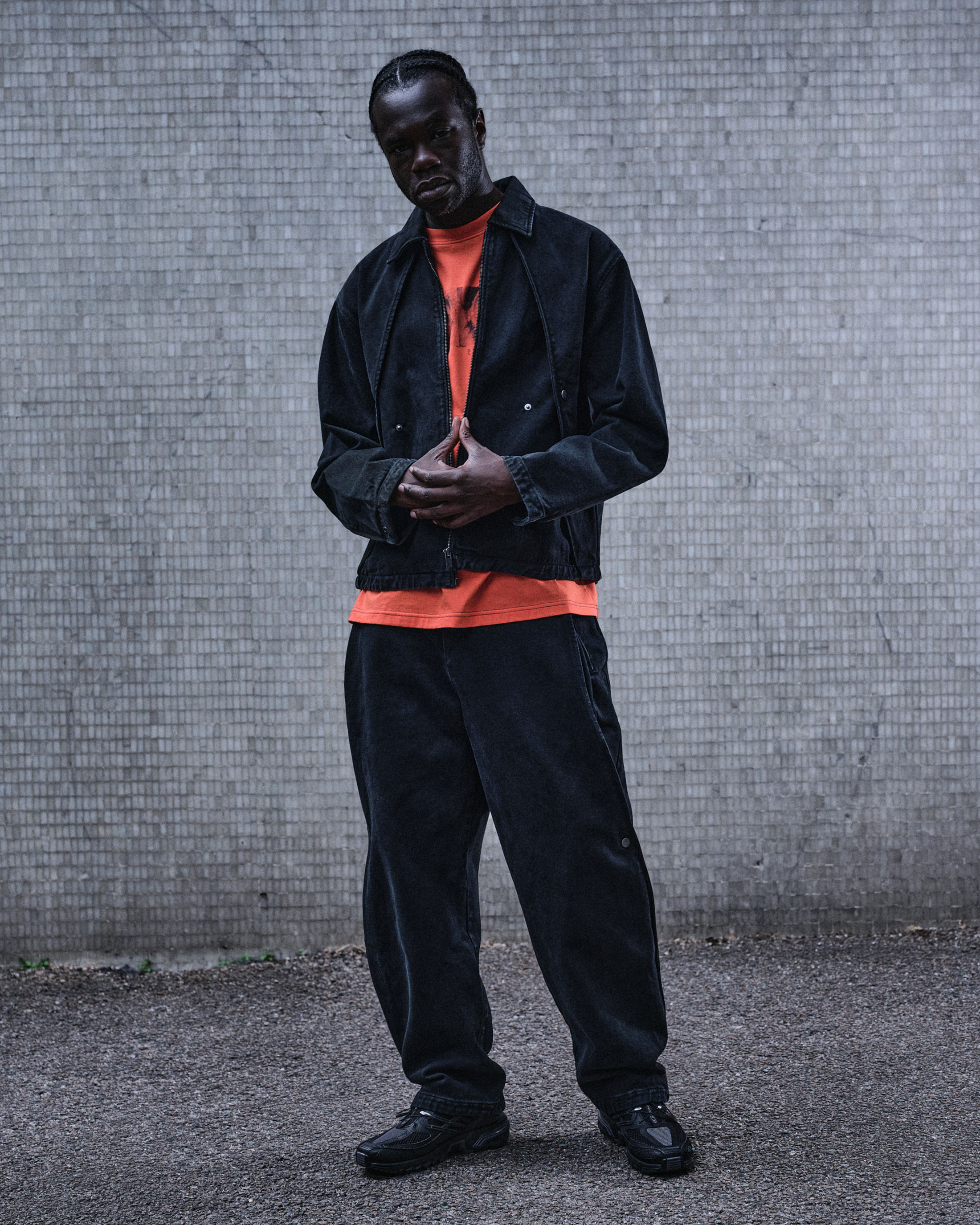 Model wearing a black jacket and matching trousers with an orange t-shirt standing against a grey concrete wall