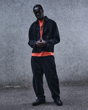 Model wearing a black jacket and matching trousers with an orange t-shirt standing against a grey concrete wall