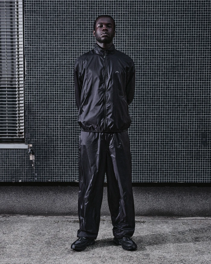 Black man standing straight against a tiled building wall wearing a dark track suit and sneakers