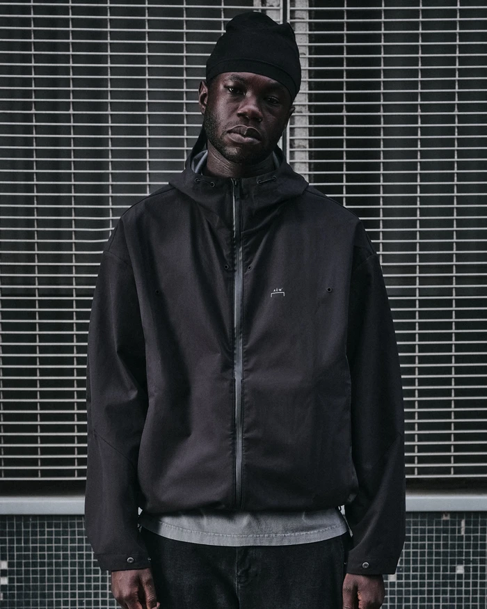 Black man wearing a dark technical jacket and beanie against a metal grille background for A-Cold-Wall