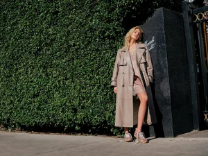 Stella Maxwell in a long camel leather trench coat leaning against a dark wall with a green hedge background.