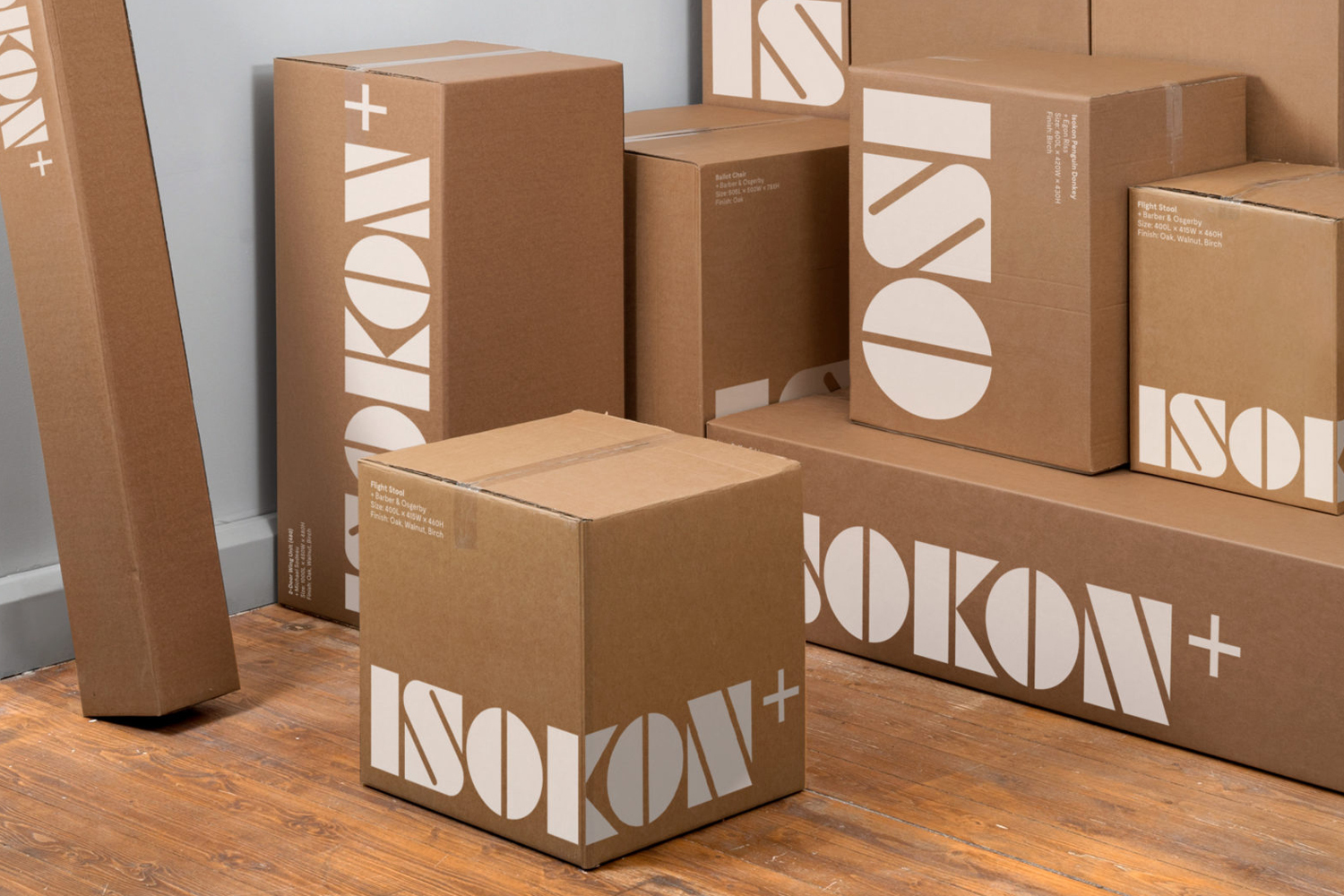 Stacked cardboard shipping boxes with bold white Isokon Plus typography on a wooden floor against a pale wall.
